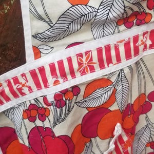 May include: Apron with a colorful fruit pattern. The apron features a cream background with large red and orange fruit illustrations, accented by black and white leaves. The apron's trim and pocket have a red and white striped design.