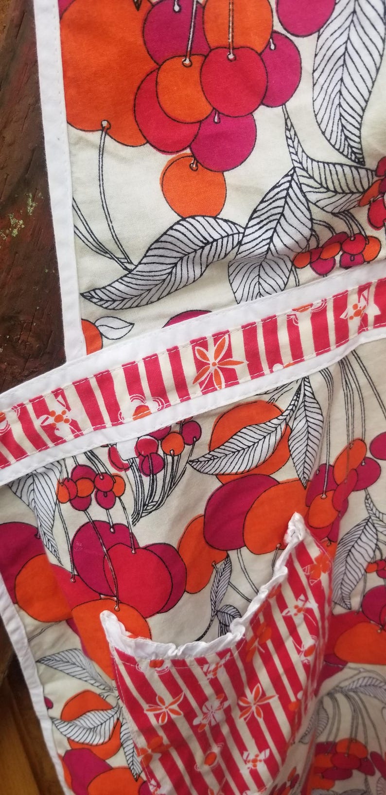 May include: Apron with a colorful fruit pattern. The apron features a cream background with large red and orange fruit illustrations, accented by black and white leaves. The apron's trim and pocket have a red and white striped design.