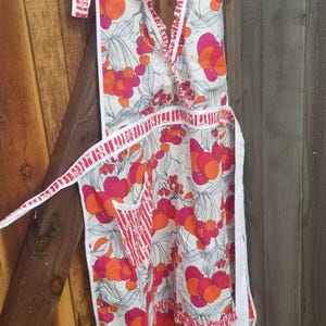 May include: A vintage-style apron with a white background and a vibrant cherry pattern in shades of red and orange. The apron features a halter neck, a pocket, and white trim. It is hanging on a black hanger.