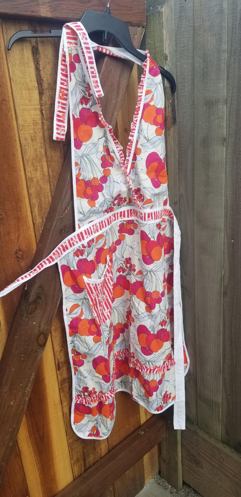 May include: A vintage-style apron with a white background and a vibrant cherry pattern in shades of red and orange. The apron features a halter neck, a pocket, and white trim. It is hanging on a black hanger.