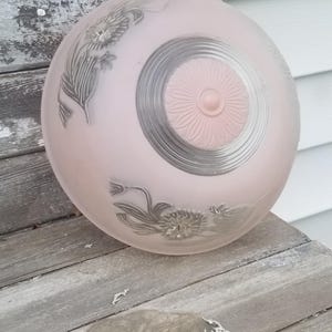 May include: A vintage, round, pink glass light fixture shade. The shade features a floral design in a darker gray color. The center has a circular design with a raised, flower-like detail. The shade is resting on a weathered wooden surface.