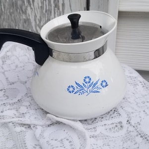 May include: White coffee pot with a black handle and lid. The pot has a blue floral design and a silver band around the middle. It sits on a white lace doily.
