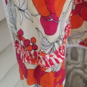 May include: Close-up of a fabric with a floral pattern. The design features large, stylized red and orange fruit shapes with black outlines on a cream background. The fabric has a ruffled edge.