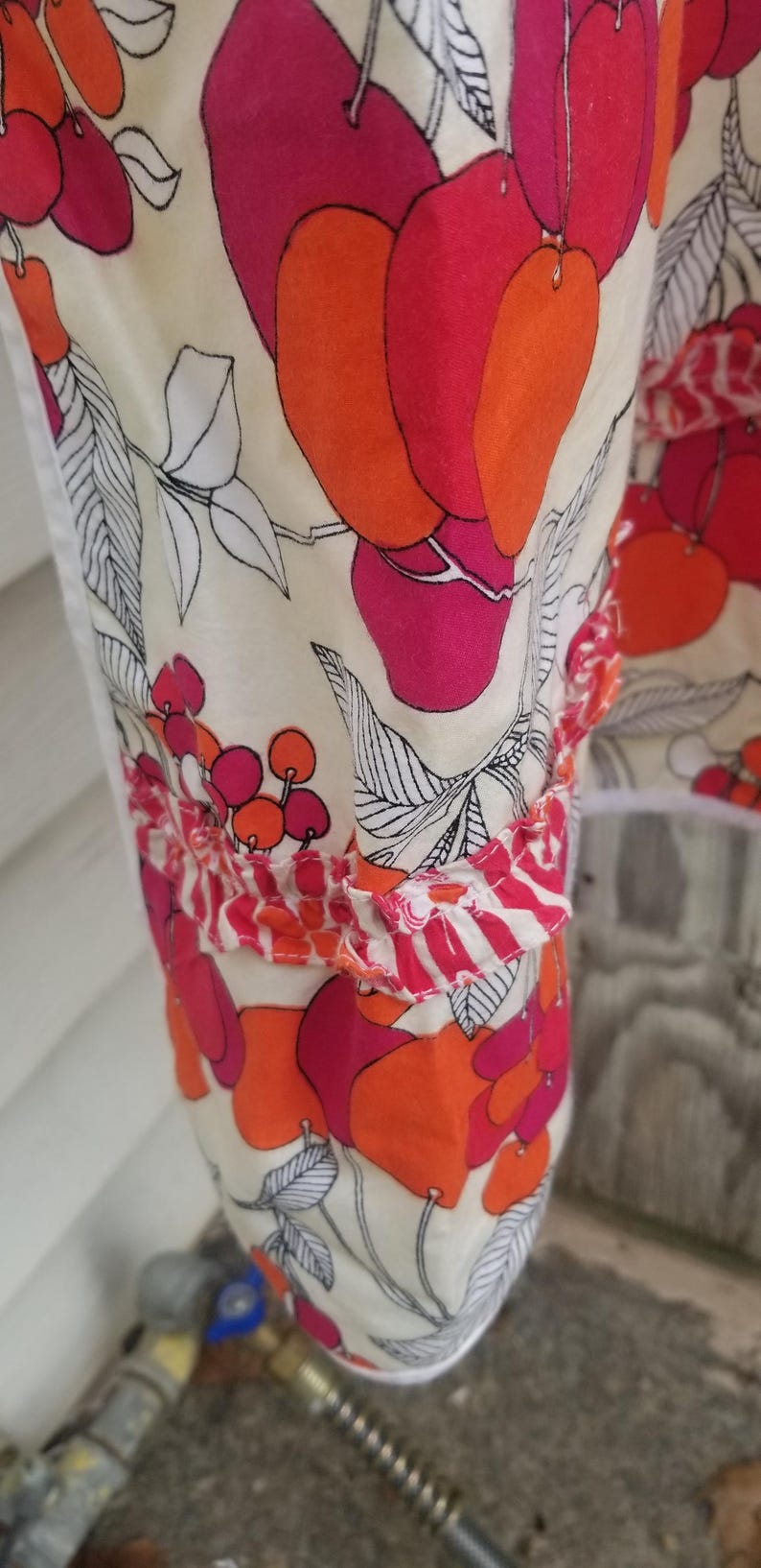 May include: Close-up of a fabric with a floral pattern. The design features large, stylized red and orange fruit shapes with black outlines on a cream background. The fabric has a ruffled edge.