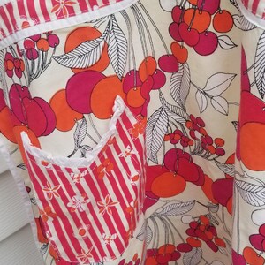 May include: Apron with a vibrant cherry pattern in shades of red and orange on a cream background. The apron features a striped pocket and trim, with white accents. The design includes black and white leaves.