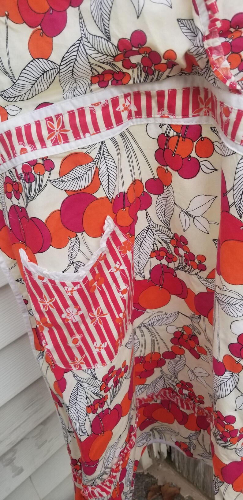 May include: Apron with a vibrant cherry pattern in shades of red and orange on a cream background. The apron features a striped pocket and trim, with white accents. The design includes black and white leaves.