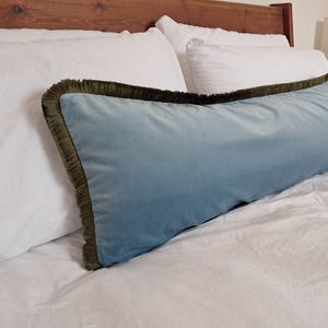 May include: A long, rectangular blue velvet cushion with a dark green fringe trim, placed on a white bed. The cushion has a soft blue hue and is set on white bedding. The headboard is made of wood.