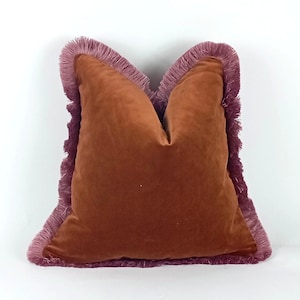 May include: A square, russet velvet cushion with a mauve fringe trim. The cushion has a soft, plush appearance and is likely used for decorative purposes. The fringe adds a touch of elegance and texture to the design.