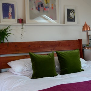 May include: A bedroom with a wooden bed frame, white bedding, and two green velvet pillows. There are two white nightstands with gold handles, one with a copper lamp and the other with a crystal ball. The walls are white and there is a white shelf with artwork above the bed.