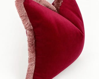May include: A square throw pillow made of deep red velvet fabric. The edges of the pillow are finished with a thick, dusty pink fringe trim that runs along the sides, providing a textured contrast to the smooth, solid-colored velvet surface.