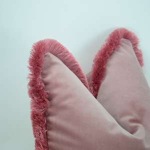 May include: Two pink velvet throw pillows with a fringe trim. The pillows are arranged side by side with the fringe trim facing outward.