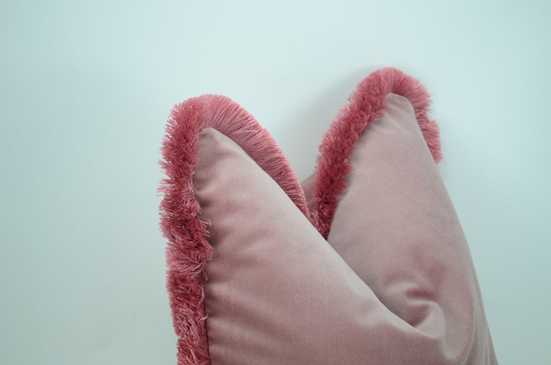 May include: Two pink velvet throw pillows with a fringe trim. The pillows are arranged side by side with the fringe trim facing outward.