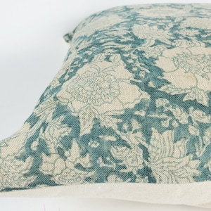 Op de afbeelding: Een vierkant decoratief kussen met een bloemenpatroon. Het kussen heeft een ontwerp van grote bloemen en bladeren in teal en gebroken wit. Het kussen heeft een neutrale, gebroken witte basis.