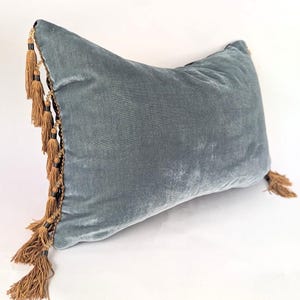 Puede incluir: Un cojín rectangular de terciopelo azul polvoriento con ribete decorativo de flecos y borlas doradas. El cojín tiene un aspecto suave y afelpado, y las borlas son de un color cálido y contrastante.