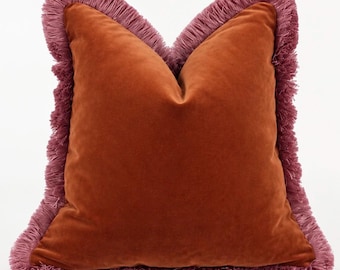 May include: A square throw pillow featuring a smooth, burnt orange velvet fabric on the front and back. The edges of the pillow are finished with a thick, textured fringe trim in a dusty mauve color that frames the entire perimeter.