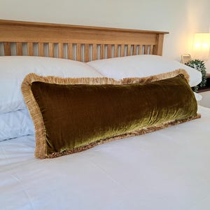 Puede incluir: Un largo cojín rectangular de terciopelo verde oliva con un ribete de flecos dorados descansa sobre una cama blanca. El cojín se coloca sobre ropa de cama blanca, con dos almohadas blancas detrás. Un cabecero de madera está en el fondo. El cojín mide unos 70 cm de largo.