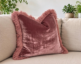 May include: A square throw pillow made of dusty rose velvet fabric. The pillow features a thick, matching fringe trim along all four edges, creating a textured border around the smooth, reflective surface of the velvet material.