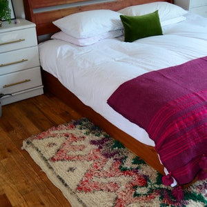 May include: A wooden bed frame with a white duvet and a burgundy throw blanket. The bed has two white pillows and a green pillow. A patterned rug is on the floor in front of the bed.