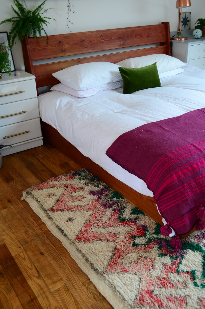 May include: A wooden bed frame with a white duvet and a burgundy throw blanket. The bed has two white pillows and a green pillow. A patterned rug is on the floor in front of the bed.