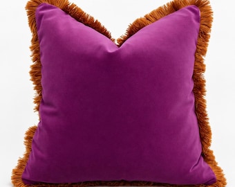 May include: A square throw pillow in a vibrant purple color featuring a smooth, solid-colored fabric front. The edges of the pillow are finished with a thick, textured fringe trim in a contrasting burnt orange or copper tone.