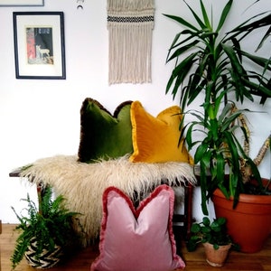May include: Three decorative throw pillows with fringed edges. The pillows are green, yellow, and pink velvet. The pillows are arranged on a bench with a fluffy white sheepskin throw.