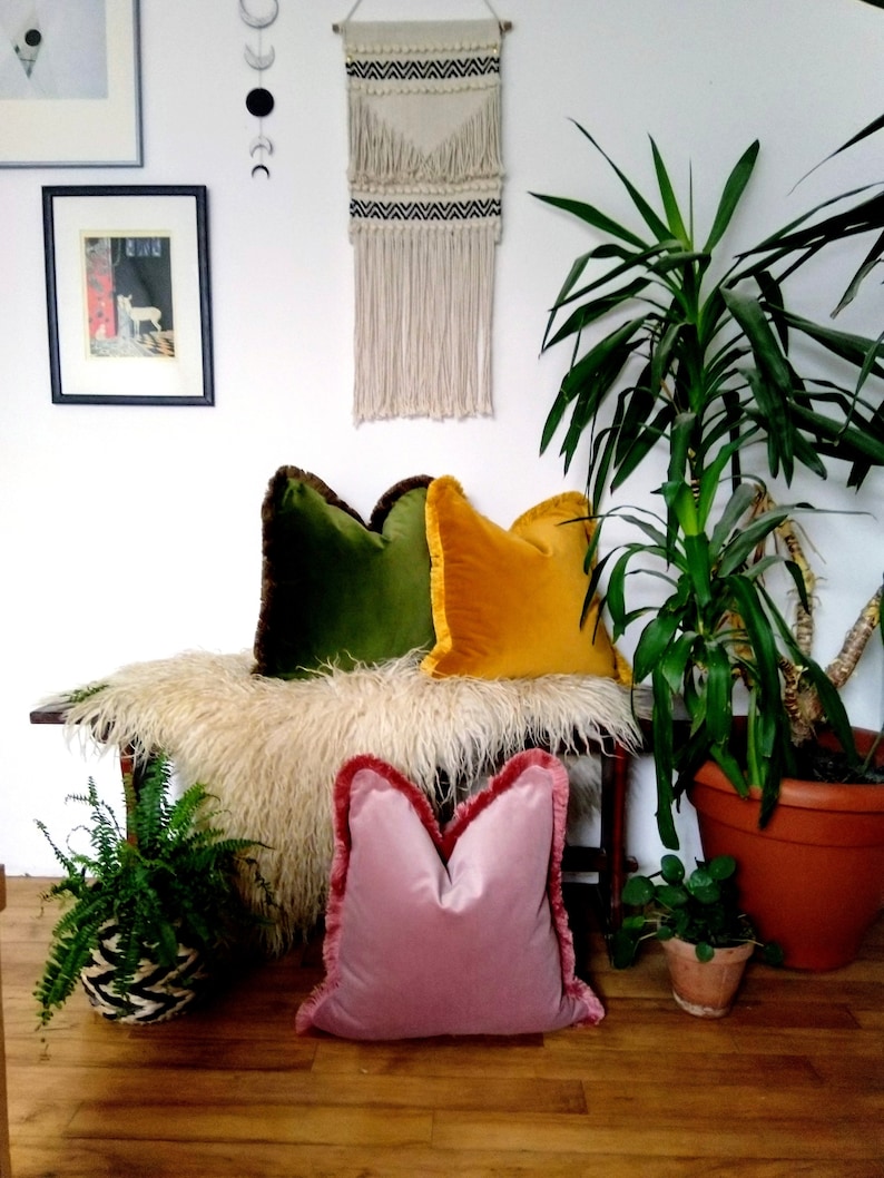 May include: Three decorative throw pillows with fringed edges. The pillows are green, yellow, and pink velvet. The pillows are arranged on a bench with a fluffy white sheepskin throw.