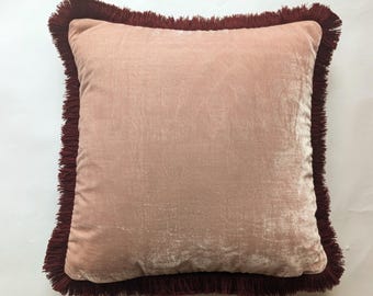 May include: A square, blush pink velvet pillow with a dark burgundy fringe trim. The pillow has a soft, textured appearance and a simple, elegant design. This decorative pillow is suitable for a bed or sofa.