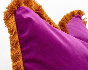 May include: A vibrant magenta velvet throw pillow featuring a thick, textured mustard yellow fringe trim along its edges. The fabric has a smooth, soft appearance, and the fringe adds a dense, layered detail to the pillow's perimeter.
