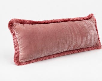 Puede incluir: Un cojín rectangular cubierto de tela de terciopelo rosa polvoriento. Los bordes del cojín están acabados con un fleco a juego que recorre todo el perímetro, creando un borde texturizado alrededor de la superficie de terciopelo liso.