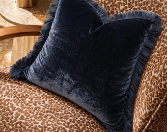 May include: A square navy blue velvet throw pillow featuring a matching fringe trim along all four edges. The pillow is positioned on a chair upholstered in a brown and tan animal print fabric with curved wooden armrests and a smooth texture.
