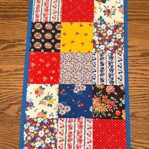 May include: A rectangular patchwork table runner with a blue polka dot border. The runner features a variety of fabric squares in red, yellow, blue, and white, with floral and polka dot patterns. The runner is placed on a wooden surface.