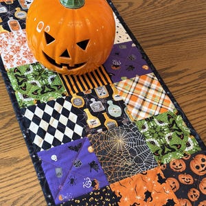 May include: A rectangular patchwork table runner featuring various Halloween-themed fabric squares with patterns of pumpkins, spiders, bats, and potion bottles, topped with a glossy orange ceramic jack-o'-lantern with a green stem.
