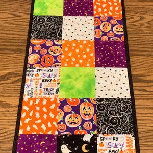 May include: A Halloween-themed table runner with a patchwork design. The runner features squares of orange, purple, green, and black fabric with Halloween motifs like pumpkins, ghosts, and spiderwebs. The border is black.