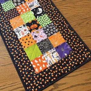 May include: A rectangular quilted table runner featuring a patchwork design of various Halloween-themed fabrics in orange, black, green, and purple. The center squares display pumpkins, ghosts, spiders, and text, bordered by a black candy corn print.