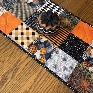 May include: A quilted table runner featuring a patchwork design of orange, black, and white fabrics with patterns of spiderwebs, pumpkins, and checkered prints, topped with a small black, orange, and white glittery decorative pumpkin ornament.