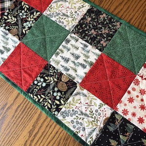 May include: A rectangular quilted table runner featuring a patchwork design of squares in red, green, black, and white. The fabric patterns include pine trees, pinecones, foliage, and snowflakes, all finished with a dark green border.