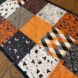 May include: A rectangular quilted table runner featuring a patchwork design of various Halloween-themed fabrics in orange, black, and white. The squares display patterns of pumpkins, black cats, witch hats, moons, stars, and spiderwebs.