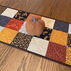 May include: A rectangular quilted table runner featuring a patchwork pattern of squares in navy blue, orange, mustard yellow, and cream with floral and geometric prints. A textured, brown, plush pumpkin with a blue stem sits on the runner.