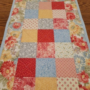 May include: A quilted table runner with a patchwork design. The center features squares of red, blue, yellow, and gray floral patterns. The border has a floral print with red, pink, and yellow flowers on a white background. A light blue border frames the runner.