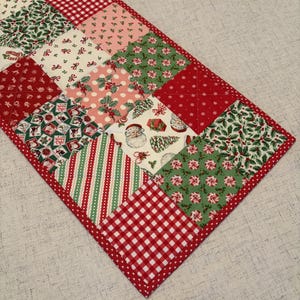 May include: A rectangular quilted table runner featuring a patchwork design of various Christmas-themed fabrics in red, green, and white. The patterns include Santa Claus faces, holly leaves, peppermint candies, red gingham, and snowflakes with a red border.