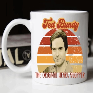 May include: White ceramic mug featuring a portrait of Ted Bundy with the text "Ted Bundy" above and "The Original Heart Stopper" below. The design includes a retro sunset graphic in shades of orange and red.