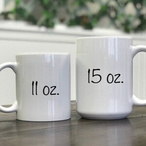 Peut inclure: Deux mugs en c&eacute;ramique blancs, l'un portant l'inscription "11 oz." et l'autre "15 oz.". Les mugs ont un design simple avec une anse incurv&eacute;e. Les mugs sont sur une surface en bois.