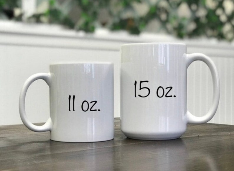 Peut inclure: Deux mugs en c&eacute;ramique blancs, l'un portant l'inscription "11 oz." et l'autre "15 oz.". Les mugs ont un design simple avec une anse incurv&eacute;e. Les mugs sont sur une surface en bois.