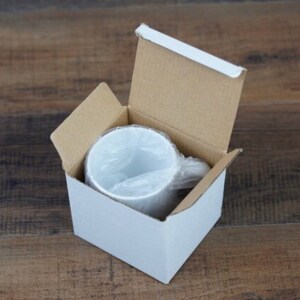 Peut inclure: Une tasse en c&eacute;ramique blanche &agrave; l'int&eacute;rieur d'une bo&icirc;te en carton blanche avec les rabats sup&eacute;rieurs ouverts. La tasse est envelopp&eacute;e dans du plastique transparent. La bo&icirc;te est sur une surface en bois.