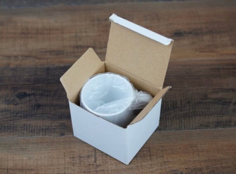 Peut inclure: Une tasse en c&eacute;ramique blanche &agrave; l'int&eacute;rieur d'une bo&icirc;te en carton blanche avec les rabats sup&eacute;rieurs ouverts. La tasse est envelopp&eacute;e dans du plastique transparent. La bo&icirc;te est sur une surface en bois.