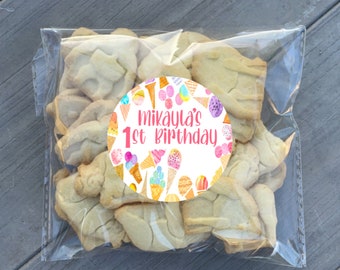 May include: A clear plastic bag filled with sugar cookies decorated with an ice cream cone design. A round sticker with a pink background and white text reads "Mikayla's 1st Birthday" and is decorated with colorful ice cream cones.