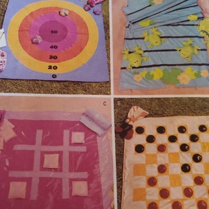 以下が含まれることがあります： 4つの異なる布製ゲームボードが掲載されたページ。ゲームには、カラフルなリングが付いたダーツボード、三目並べボード、チェッカーボード、カエルをテーマにしたブランケットとスイレンが含まれています。