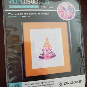 May include: A packaged cross-stitch kit labeled "Artiste Mini" with a "Wild at Heart" design. The kit includes needle, floss, and instructions. The finished design size is 4.5in x 6.13in. The image features a framed cross-stitch of a teepee in pink, purple, and orange.