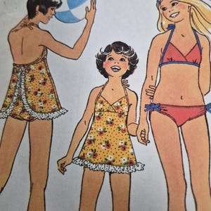 May include: A sewing pattern illustration featuring three figures wearing swimwear. One figure wears a yellow floral halter top with white ruffles and matching shorts. Another figure wears a red bikini with blue trim and side ties. A blue and white beach ball is visible.