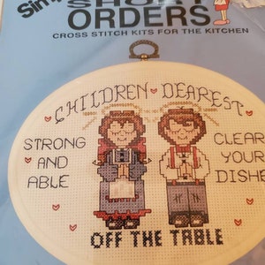 May include: A cross-stitch kit by Simplicity, featuring a design with the text "Children Dearest, Strong and Able, Clear Your Dishes, Off the Table." The kit includes a stitched oval with two figures and hearts.
